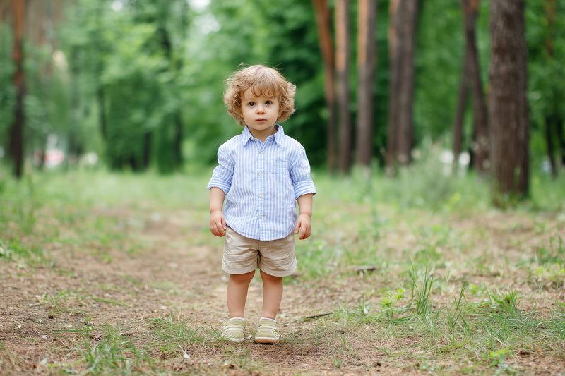 Curly haired toddler boy stands on a forest path surrounded by green trees and grass, wearing a striped shirt and shorts, looking curious and serene in soft sunlight during a summer outing.
