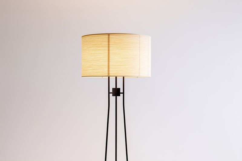 Minimal modern floor lamp with woven fabric shade casting warm ambient light against a neutral wall, creating cozy contemporary interior mood and subtle decorative accent.