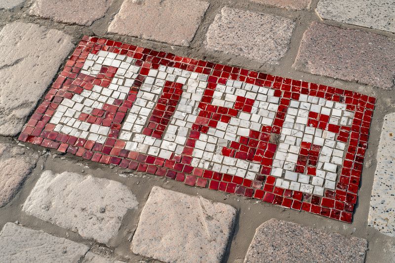 Red and white mosaic tile displaying the number 2026 embedded in a cobblestone pavement, weathered textured tiles forming numerals that contrast with the rough stone street surface.