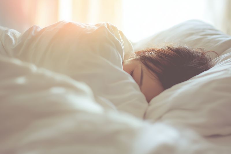 Young person sleeping peacefully under soft white blankets in a sunlit bedroom, cozy morning scene with warm natural light creating a calm and comfortable atmosphere.