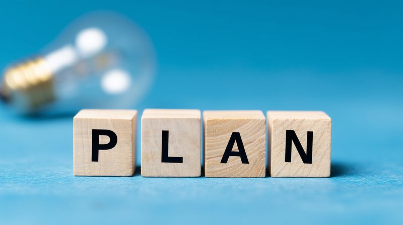 Wooden blocks spelling PLAN on a blue surface with a blurred light bulb in the background, symbolizing planning, strategy, idea generation and business preparation for projects and goals.