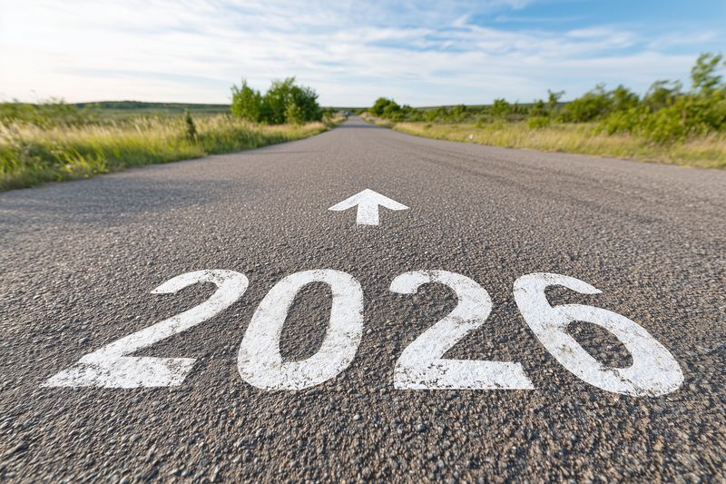 Low angle view of a paved asphalt road with bold white numerals 2026 and a forward arrow painted on the surface, stretching toward a distant horizon under a clear blue sky.