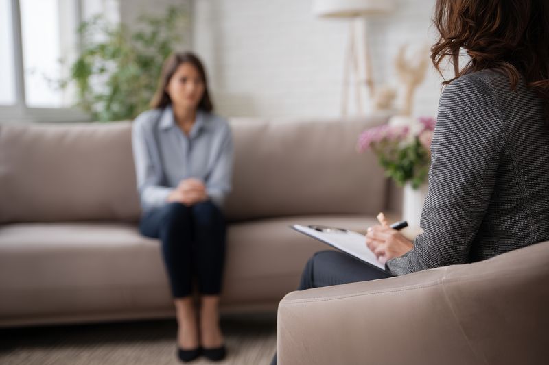 Young woman sitting on a sofa during a private therapy session while a counselor takes notes, offering a calm and supportive environment for emotional discussion and mental health care.