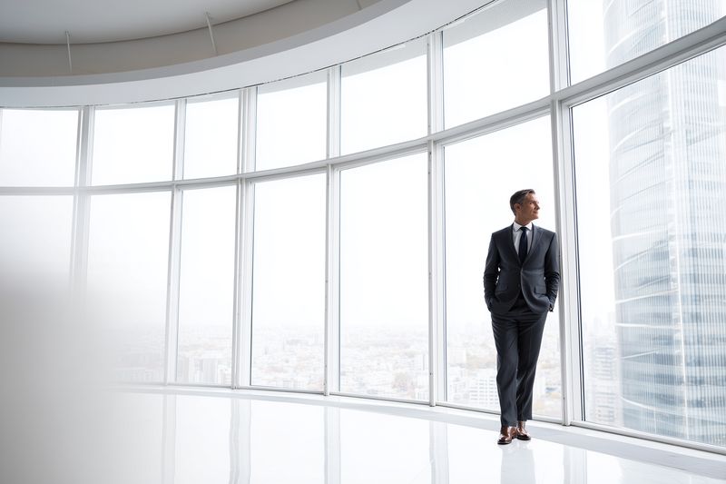 Confident businessman in a dark suit standing near expansive curved floor-to-ceiling windows in a modern highrise office, overlooking a bright cityscape through glass facade.