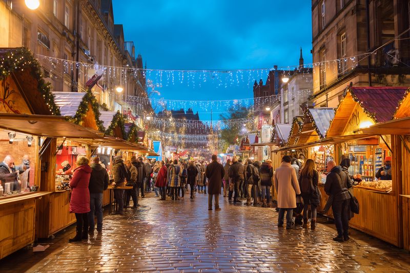 Evening Christmas market scene with wooden stalls along a wet cobblestone street, twinkling string lights overhead and crowds browsing festive gifts, food stalls and crafts.