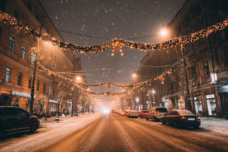 Snowy urban street at night decorated with festive string lights and garlands, warm streetlamps glowing, parked cars along the road, gentle snowfall creating cozy winter atmosphere.