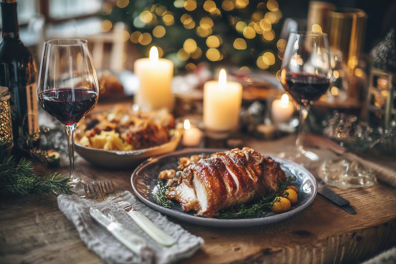 Festive holiday table setting with roasted pork loin on a plate surrounded by roasted vegetables, glowing candles, wine glasses and warm bokeh lights creating cozy atmosphere.
