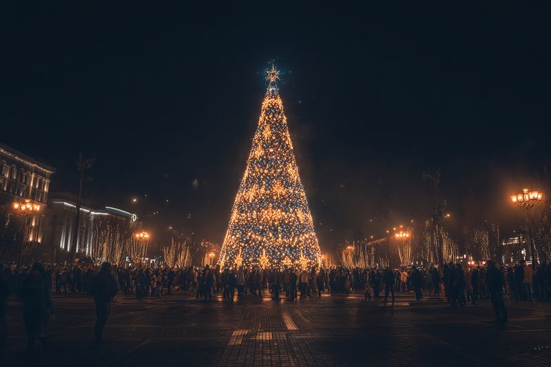 A large illuminated Christmas tree stands in a bustling city square at night, surrounded by crowds and festive lights casting warm golden glow over the holiday scene.