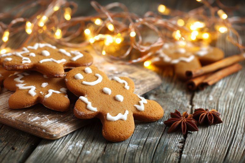 Festive gingerbread cookies with white icing arranged on a rustic wooden board surrounded by star anise, cinnamon sticks and warm fairy lights creating cozy holiday atmosphere.