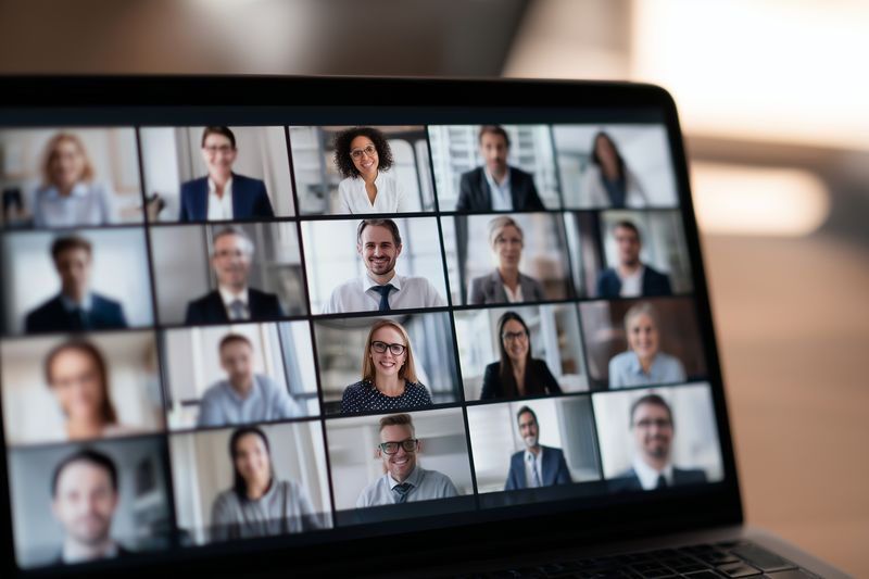 A blurred laptop screen displays a virtual video conference grid of professionals in a remote business meeting, illustrating online teamwork, communication, digital workplace interaction and