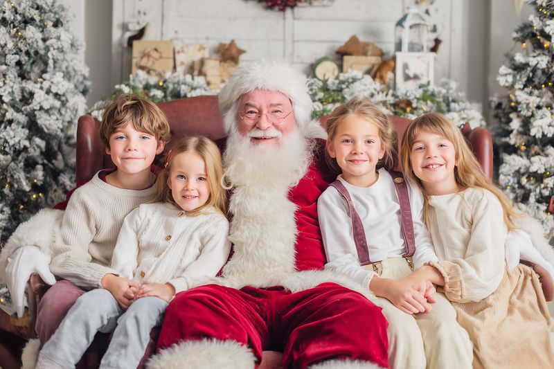 Bearded man in a festive red costume sits on a leather chair surrounded by four smiling children wearing cozy winter clothes, with holiday ornaments, wrapped gifts and decorated trees.