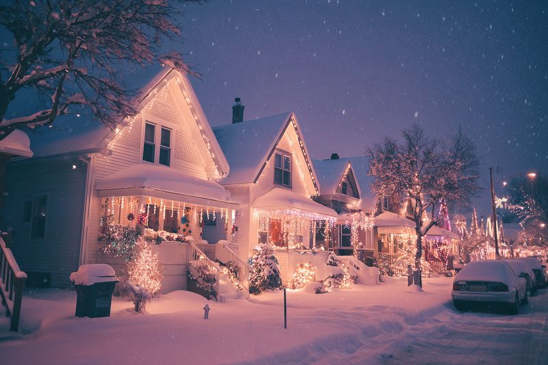 Snowy residential street at night with charming decorated houses and warm festive lights glowing through falling snow, cozy porches, snow-covered cars and trees in a serene winter neighborhood scene.