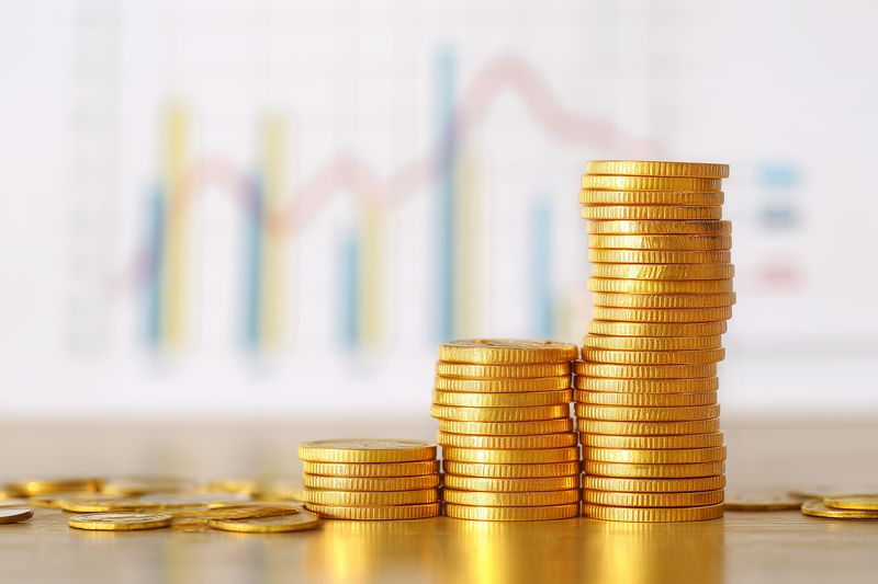 Close-up of stacked gold coins arranged in ascending columns on a reflective surface with a blurred financial chart in the background, symbolizing investment growth and economic success.