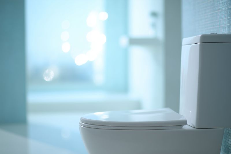 Close-up of a modern white toilet bowl and tank in a clean minimalist bathroom interior, with soft blue bokeh background and gentle natural light highlighting smooth ceramic and sanitary surfaces.