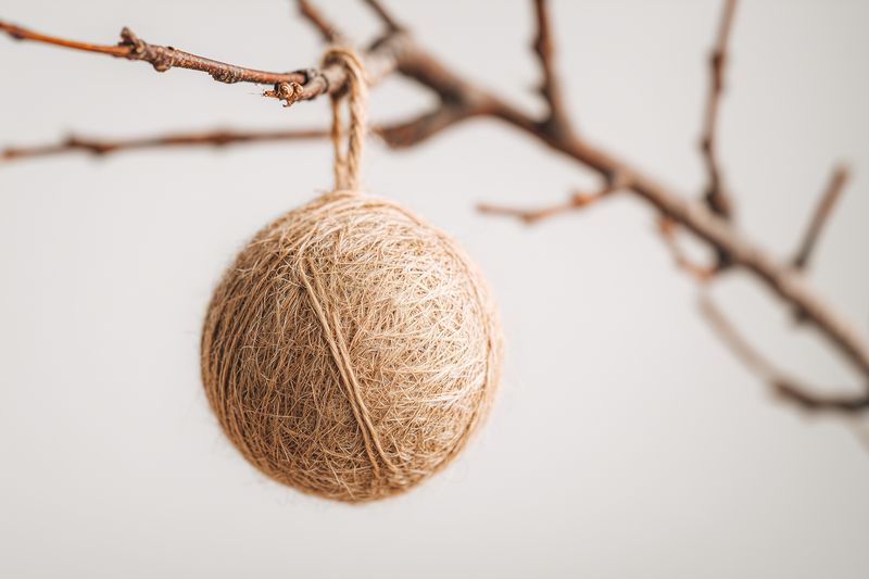 A minimalist jute twine ball ornament hanging from a bare twig, highlighting rustic texture and organic fibers against a soft neutral background for seasonal natural decor.