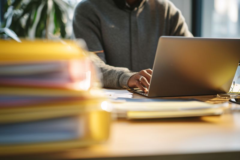 Person working on a laptop at a cluttered desk with folders and documents, soft natural light creating a warm productive atmosphere in a modern home office or coworking workspace.