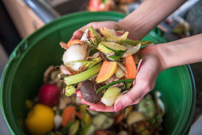 Hands holding assorted fruit and vegetable scraps over a green compost bin in a kitchen, illustrating household organic waste collection and home composting for sustainable living.