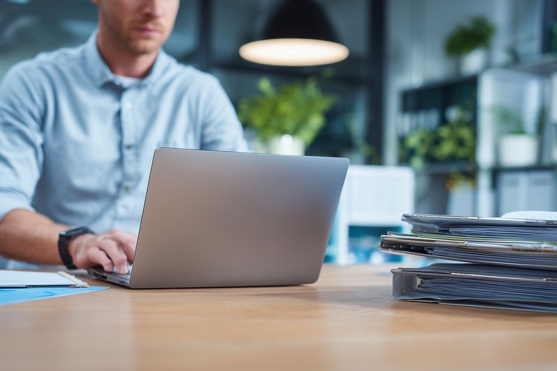 A focused professional types on a laptop at a wooden desk in a modern office environment, with neatly stacked file folders and documents waiting to be reviewed nearby.