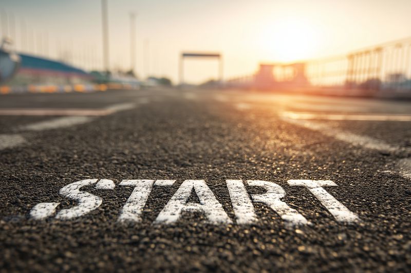 Close-up of the painted START word on asphalt at a racing track, low angle perspective with sun flare and shallow depth of field, evoking motion, anticipation and the beginning of a race.