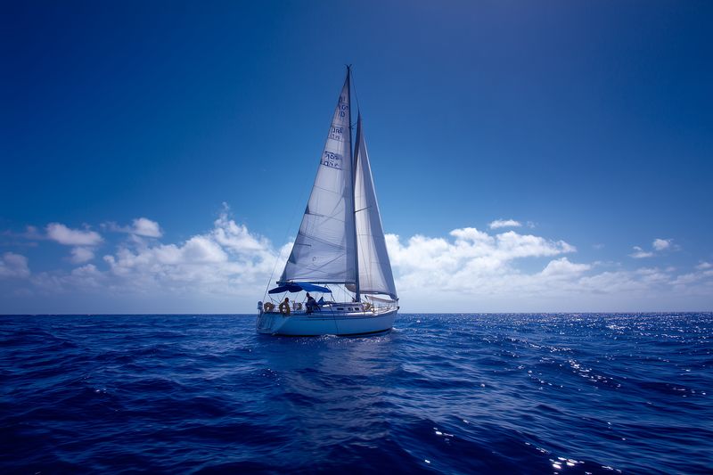 Sailboat cruising on a deep blue ocean under a clear sky, white sails filled with wind, gentle waves and bright sunlight creating a serene maritime scene for travel and leisure.