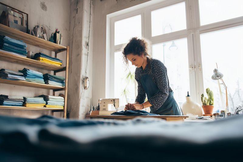 Young seamstress working at a sewing machine in a sunlit studio, surrounded by neatly folded fabrics and shelves, concentrating on handcrafted garment construction and design.