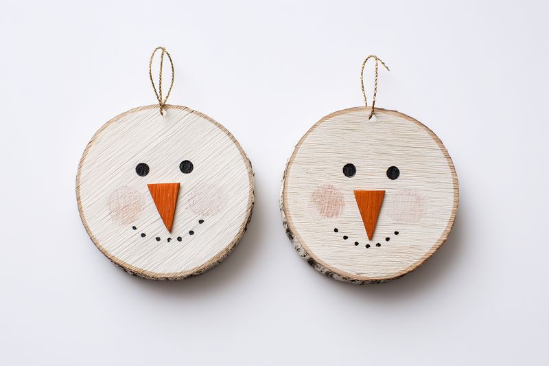Two handmade wooden holiday ornaments painted as minimalist snowman faces with carrot noses, button eyes and simple smiles, rustic craft decorations photographed on a white background.