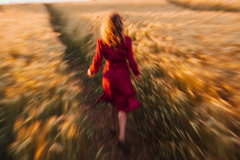Blurred motion of a woman in a red dress running barefoot along a narrow path through a golden wheat field at sunset, creating dynamic warm and dreamy countryside atmosphere.