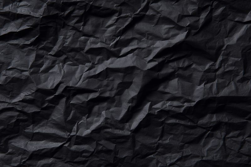 High contrast crumpled black paper texture with dramatic folds and highlights, suitable as background for graphic design, packaging mockups, and editorial layouts requiring dark textured surface.