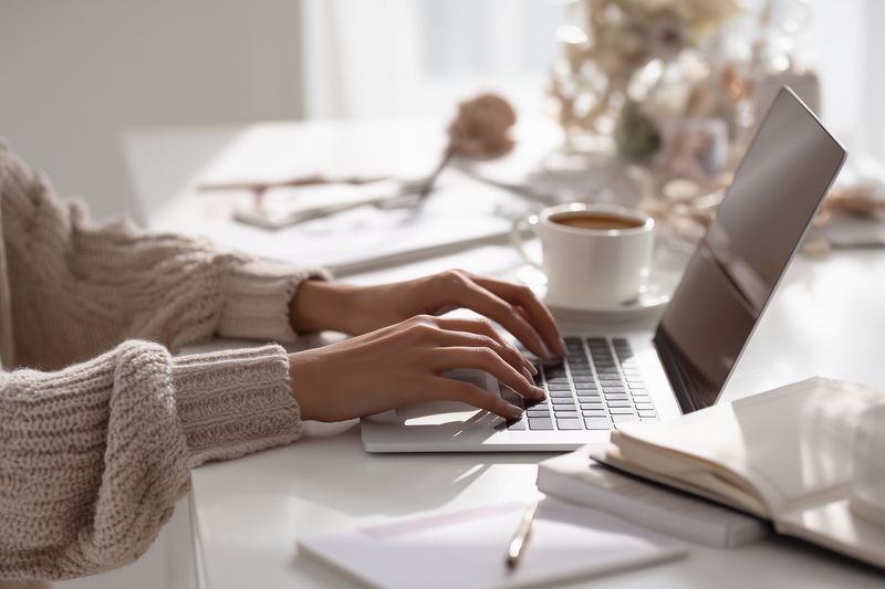 Closeup of hands typing on a laptop at a bright home desk, surrounded by a notebook, pen and a cup of coffee, evoking cozy remote work, productivity and modern lifestyle ambiance.