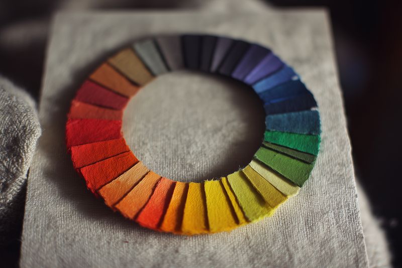 Close-up of a handcrafted color wheel made from felt swatches arranged in a circular rainbow on textured fabric, showcasing vibrant hues, tactile texture and subtle shadow details.