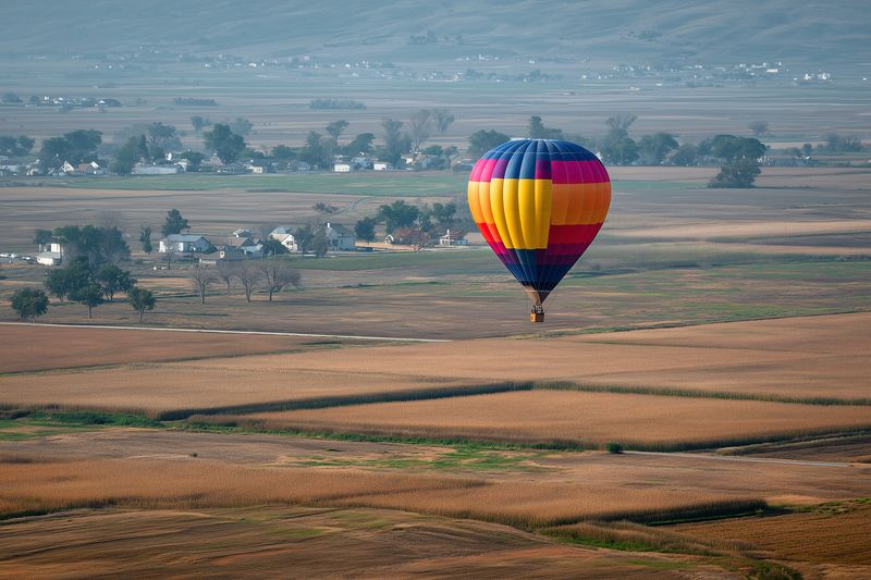 A colorful hot air balloon floats over golden farmland and patchwork fields at sunrise, creating a serene aerial landscape with scattered houses, trees and wide open rural views.