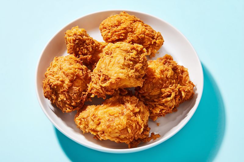 Studio photo of golden crispy fried chicken pieces on a white plate against a bright blue background, showcasing crunchy breading, warm tones, and appetizing texture in closeup.