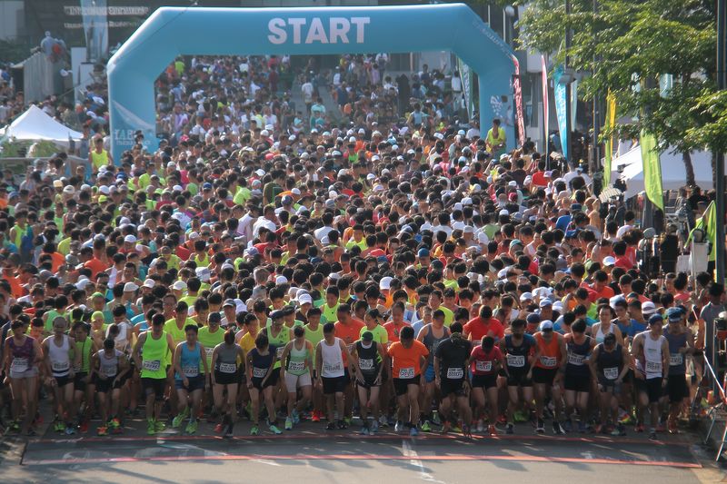 A dense pack of runners surges forward under a start arch at the beginning of a road race, capturing energy, teamwork and determination in a vibrant outdoor athletic event.