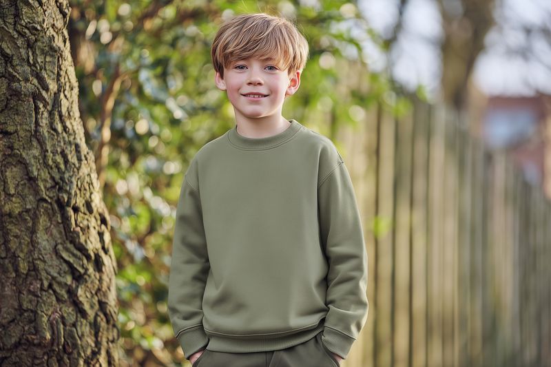 Portrait of a smiling young boy standing outdoors by a tree and wooden fence, wearing a green sweater. Natural light highlights warm skin tones and a relaxed candid expression.