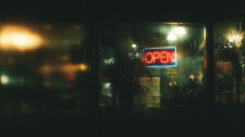 Neon open sign glowing in a rainy storefront window at night, casting warm reflections on glass and revealing silhouetted interior plants and cozy ambient lighting.