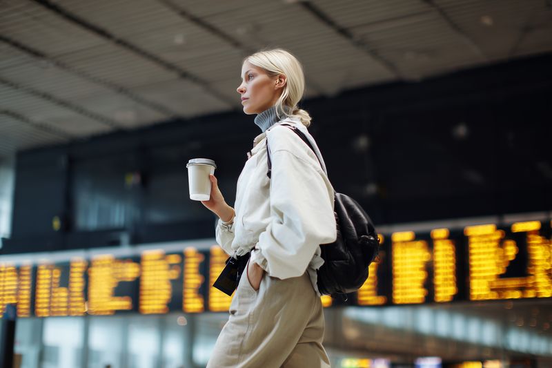 Young female traveler carrying a takeaway coffee cup and wearing a backpack walks through a bustling airport terminal, glancing up at the illuminated departure board while heading to her gate.