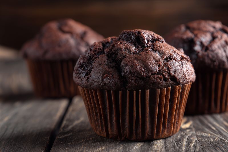 Close-up of three homemade chocolate muffins on a rustic wooden table, showcasing rich texture, crackled tops and moist crumb in warm natural light for appetizing food photography.