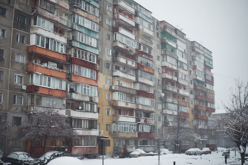 Snow falls across an aging concrete apartment block with enclosed balconies and parked cars, creating a moody urban winter scene of residential architecture and muted colors in cold weather.