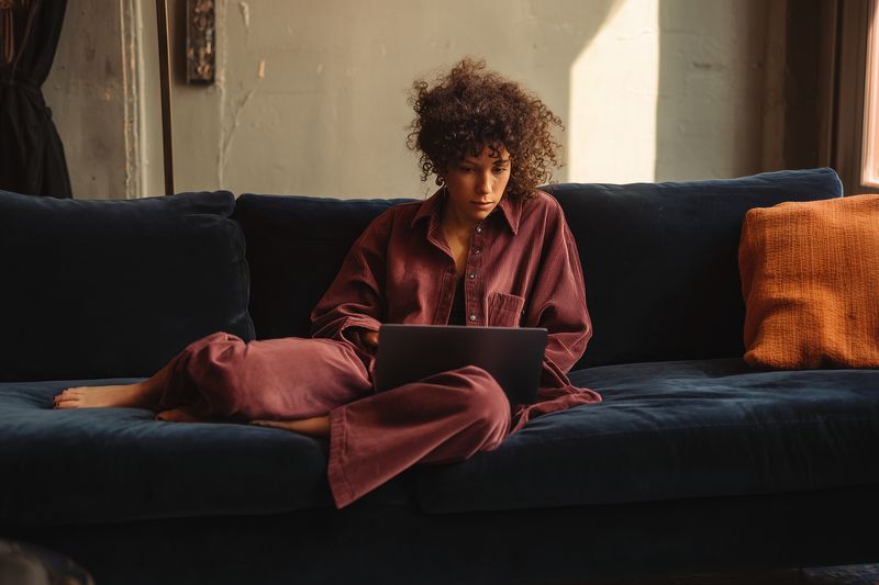 Young woman with curly hair lounges on a dark sofa using a laptop, working remotely in a cozy living room with warm natural light and relaxed casual loungewear.