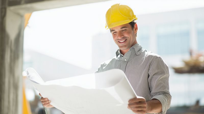 Smiling construction engineer wearing a yellow hard hat reviews architectural blueprints on site, focused on project planning and structural details in a bright urban building environment.