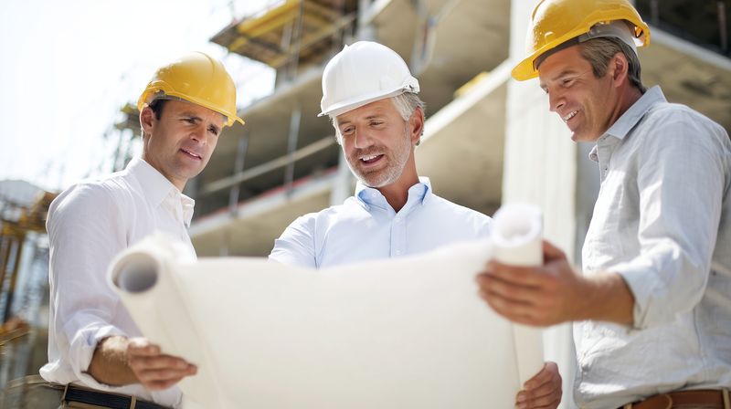 Three construction professionals wearing hard hats examine architectural blueprints at an active building site, collaborating on plans, safety, and project progress under daylight.