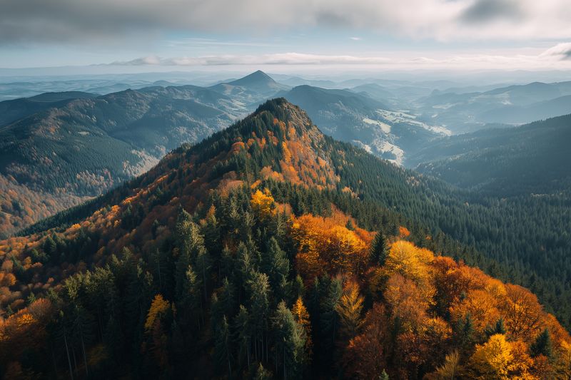 Aerial view of a forested mountain ridge in autumn with colorful foliage, misty valleys and distant peaks under dramatic clouds, capturing serene wilderness and panoramic natural beauty.