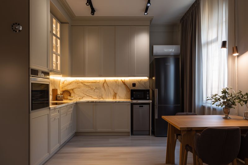 Cozy modern kitchen interior with warm undercabinet lighting, marble backsplash, integrated appliances, wooden dining table and soft curtains creating inviting cozy home space.