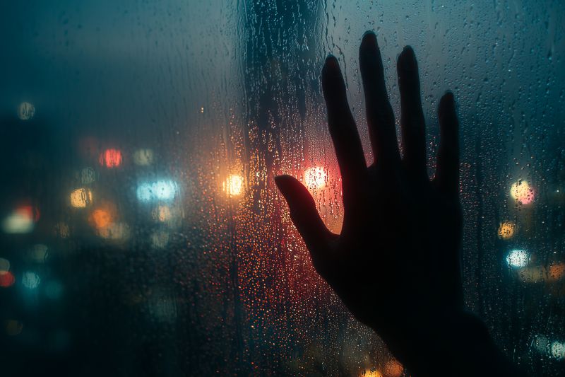 Hand silhouette on rainy window with raindrops and blurred city lights bokeh at night, moody urban scene with reflection on wet glass, cinematic glow and soft focus atmosphere