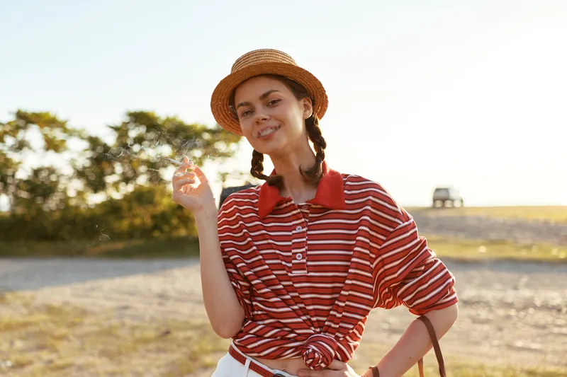 Young woman enjoying lifestyle outdoors in a striped shirt and hat. She smiles while holding small flowers in nature. Sunny rural landscape background for leisure and summer travel.