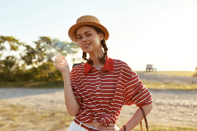 Young woman in a straw hat and striped shirt smiles while holding a cigarette outdoors, capturing an authentic lifestyle moment during a summer vacation trip.