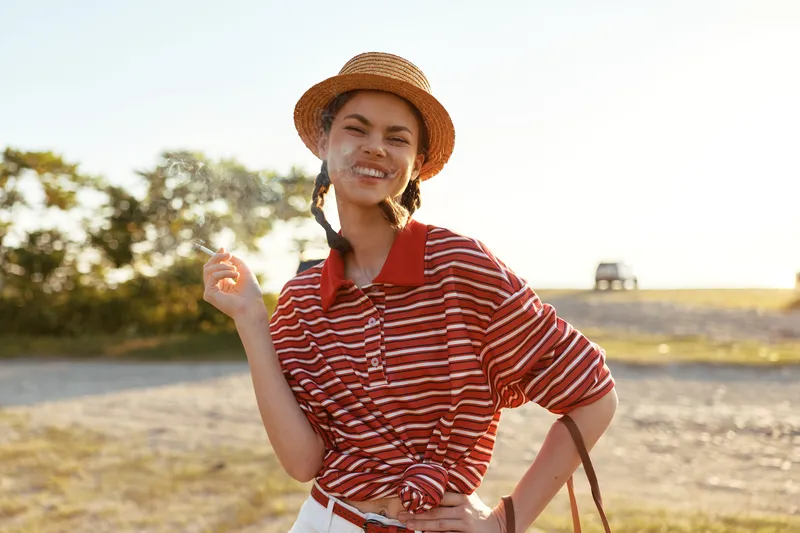 A smiling young woman in a straw hat holding a cigarette outdoors in nature. A lifestyle portrait reflecting casual leisure and freedom during a sunny summer day in sunlight.