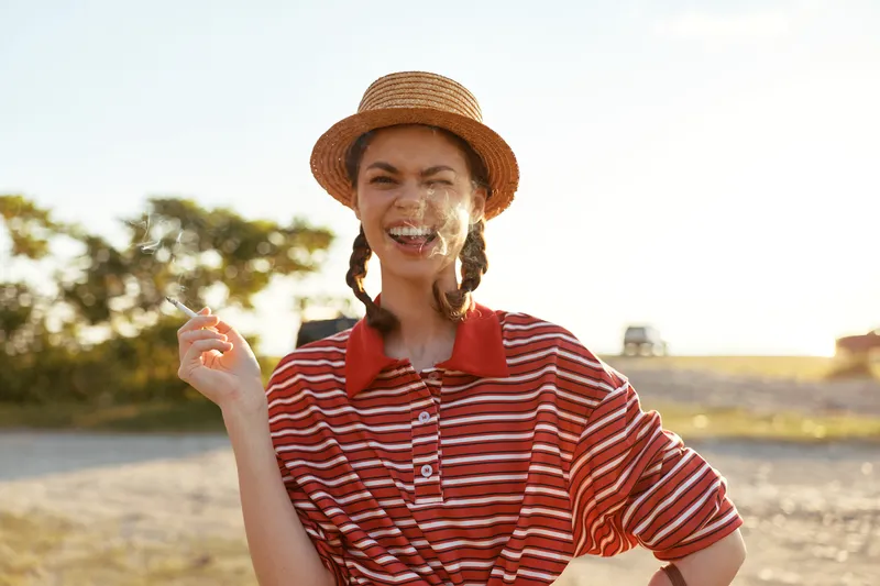 A joyful young woman with braided hair and a straw hat smokes a cigarette outdoors. The authentic lifestyle scene captures a relaxed moment of freedom in a sunny summer, creating a vibrant mood.
