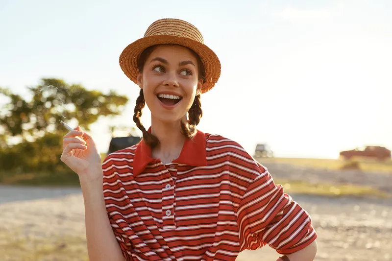 Smiling woman holding cigarette outdoors. Stylish lady wearing straw hat and striped shirt. Lifestyle portrait showing cheerful emotion during sunny day at the beach in summer season.