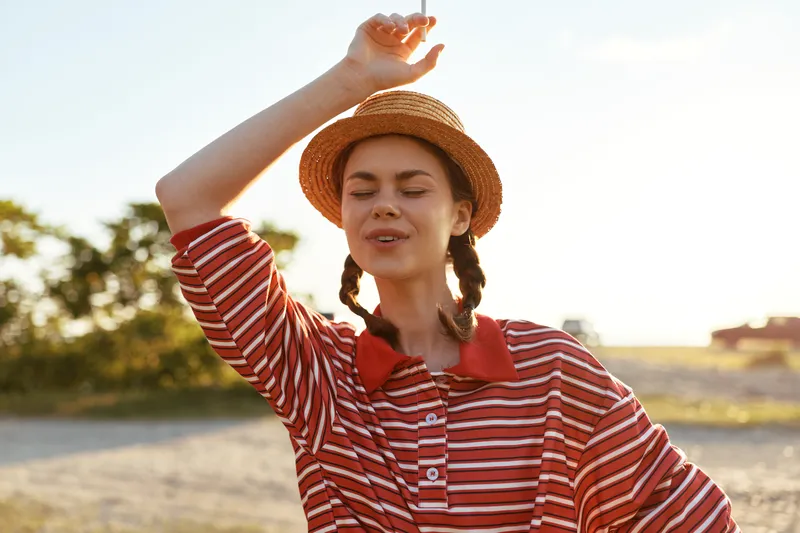 Young woman enjoying lifestyle in countryside. She wears straw hat and striped shirt, posing outdoors under sunlight. Concept for summer vacation, happiness, freedom and rural leisure time.